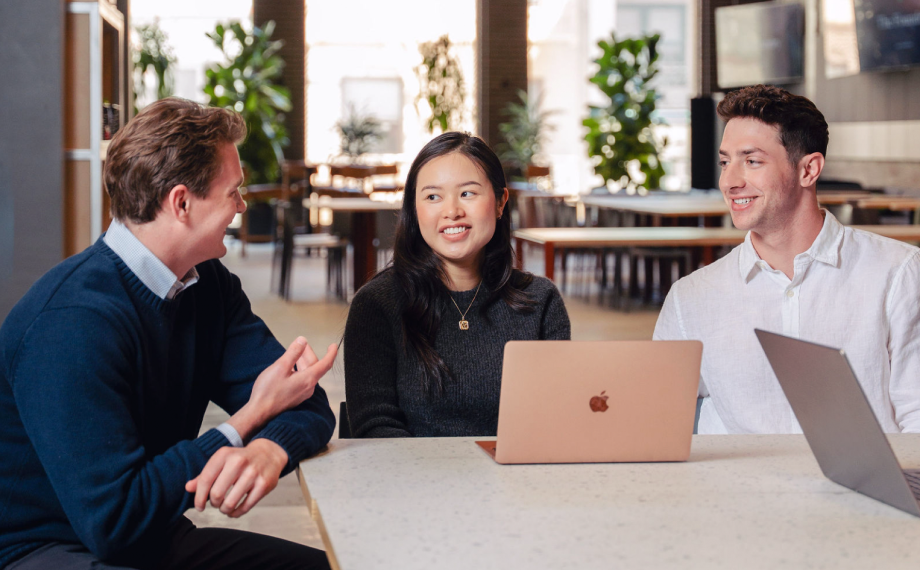 Three young adults sit at a table with laptops, smiling and talking in a modern, well-lit workspace with large windows and greenery in the background.