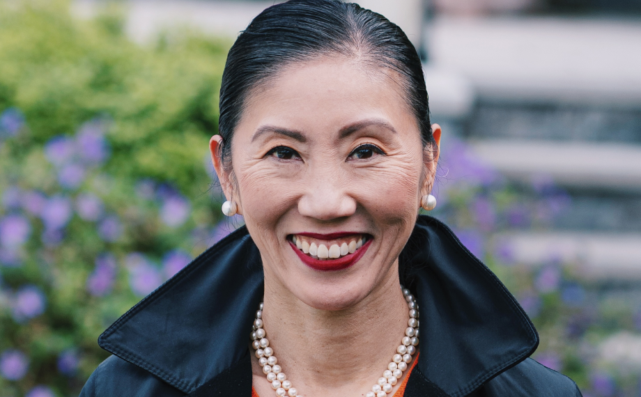 A woman with dark hair pulled back, wearing pearl earrings, a pearl necklace, red lipstick, and a dark jacket smiles outdoors with greenery and purple flowers in the background.