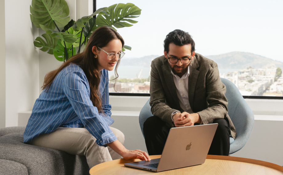 Two people sit in a modern office, looking at a laptop on a round wooden table. One points at the screen while discussing ai for career growth as the other watches. A large window and green plant are in the background.