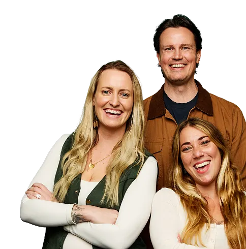 Three smiling adults pose together, two women in front with long hair and a man in a brown jacket standing behind them. All look cheerful and relaxed against a plain background.